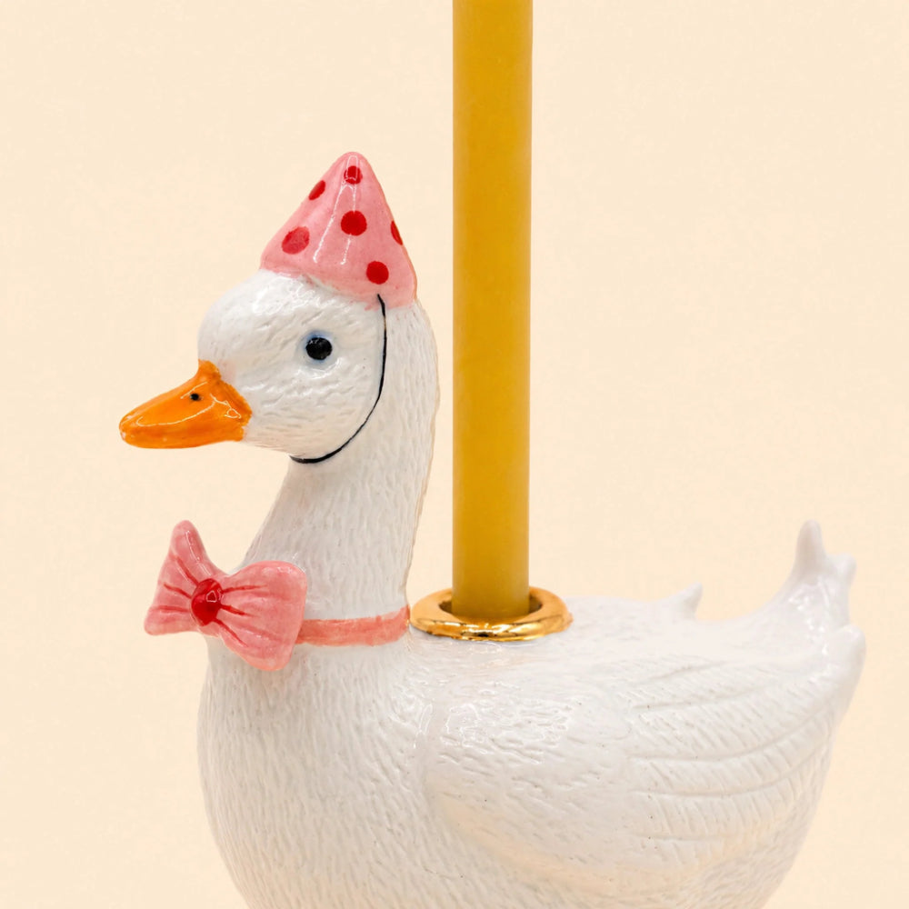 A white duck figurine with an orange beak, wearing a pink party hat with red polka dots and a pink bow tie, with a yellow candle inserted into its neck, set against a solid pale beige background.