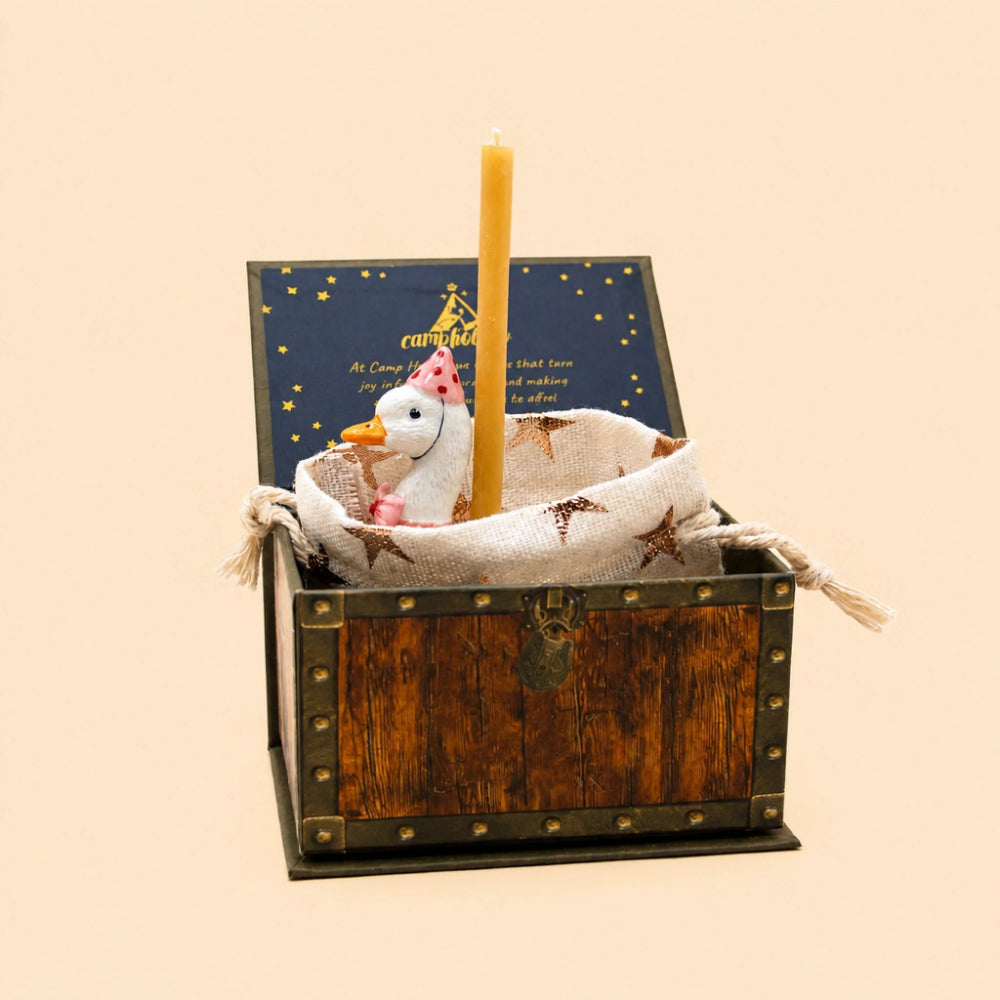 A decorative wooden chest with brass-style corner rivets and a latch, shown open on a plain beige background. Inside the chest is a burlap sack holding a white plush snowman wearing a pink party hat, with a tall yellow candle standing upright and a dark blue printed insert leaning behind it.