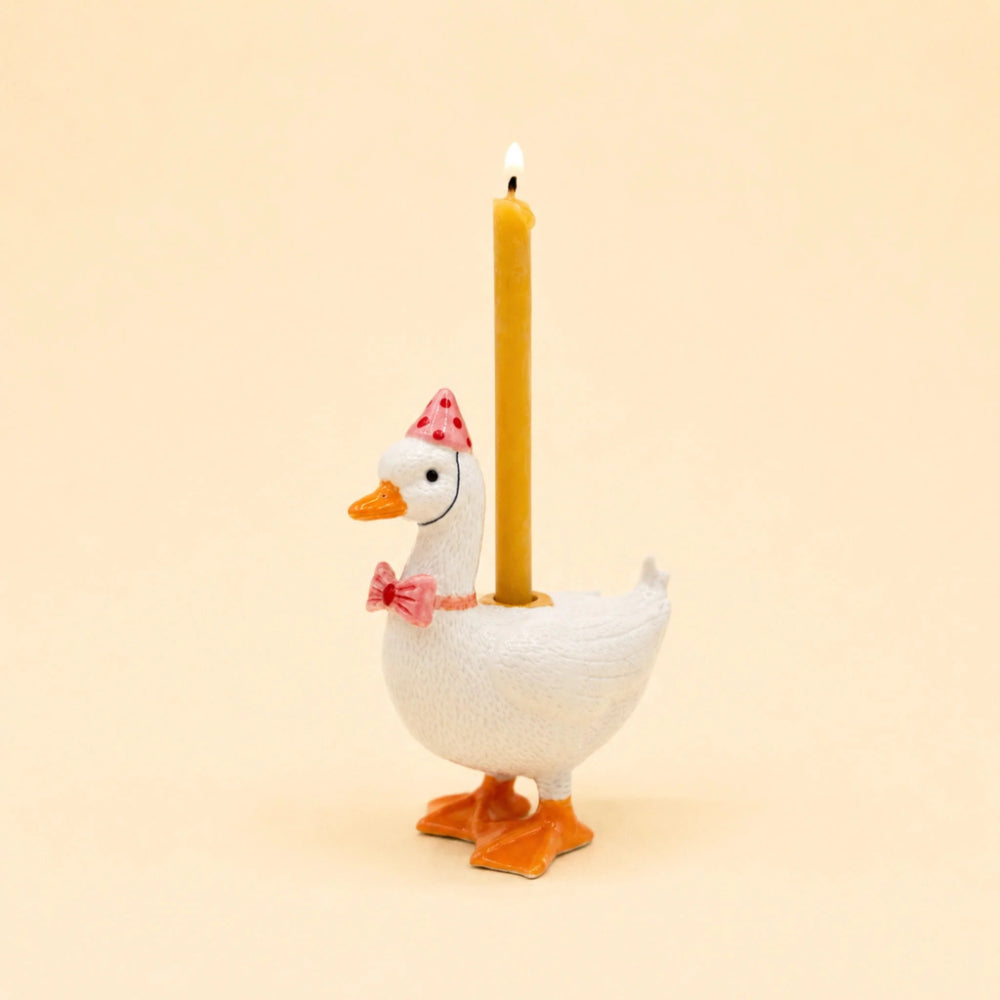 A white goose-shaped candle holder with orange feet and beak, wearing a pink polka-dot party hat and pink bow tie, holding a lit yellow candle against a warm beige background.