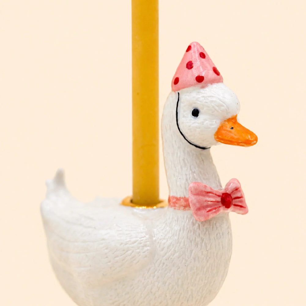 A white goose figurine with a pink polka-dot party hat, orange beak, and pink bow tie, attached to a yellow pole against a light beige background.
