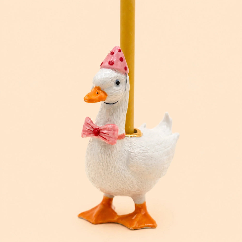 A white decorative goose figurine with orange beak and feet, wearing a pink polka-dot party hat and a pink bow tie, standing on a gold pole against a warm beige background.