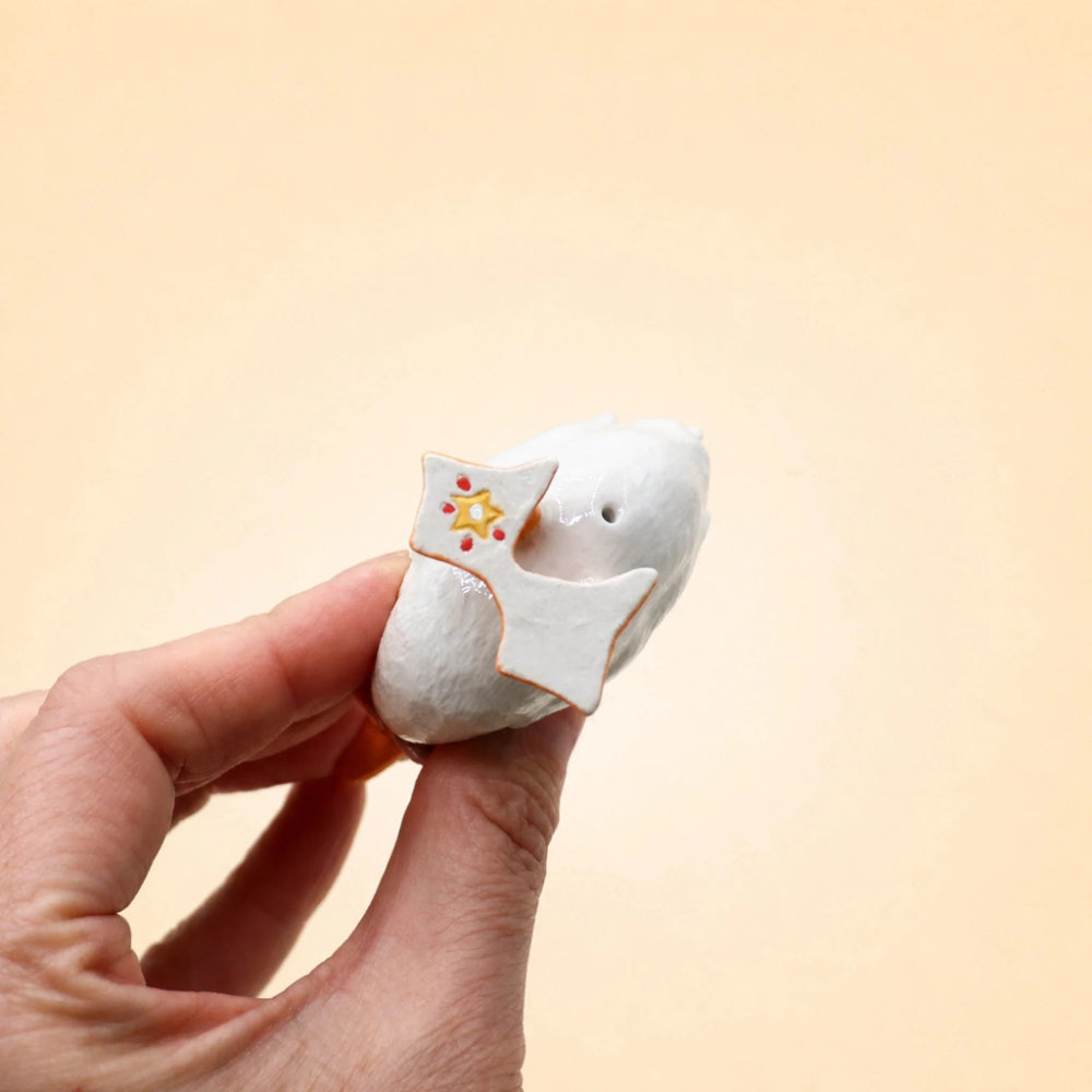 A hand holding a small white ceramic bird figurine with a star-shaped accent and tiny colorful details, set against a solid pale beige background.