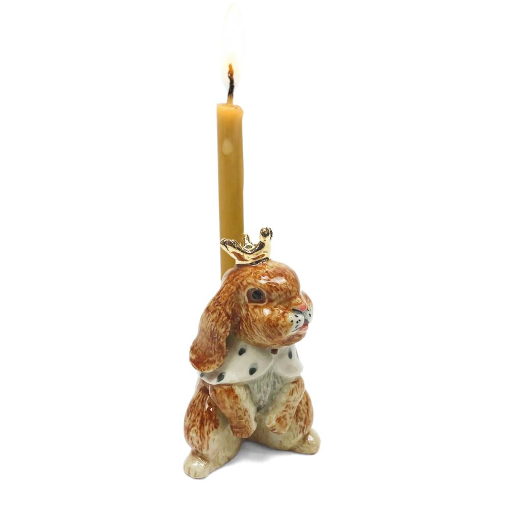 A ceramic rabbit-shaped candle holder with a lit yellow taper candle inserted, featuring a small gold crown accent on its head and a white collar with black spots, set against a plain white background.