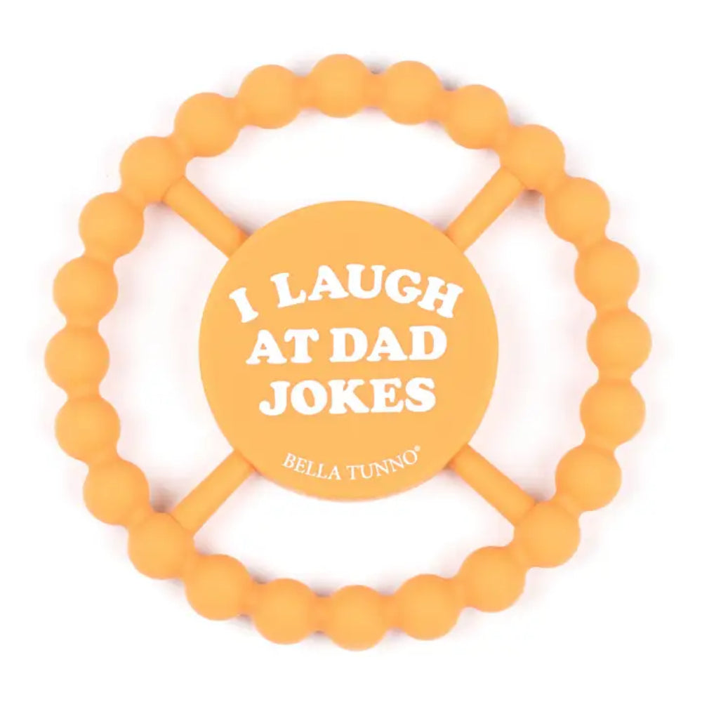 An orange silicone baby teether with a beaded outer ring and a central circular pad that reads 'I LAUGH AT DAD JOKES' with the brand name 'BELLA TUNNO' printed below in white text, on a solid white background.