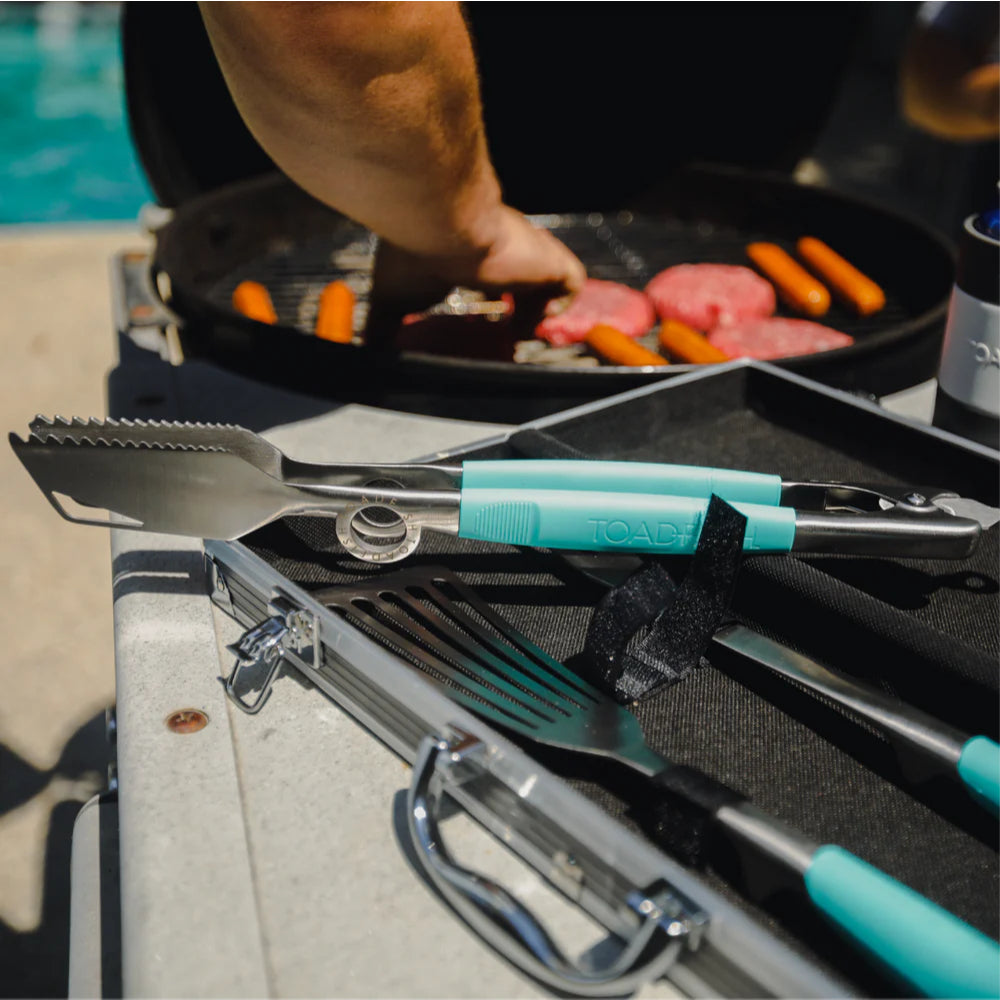 Toadfish Ultimate Grill Tongs barbecue tools with turquoise handles on a grill, with food cooking in the background.