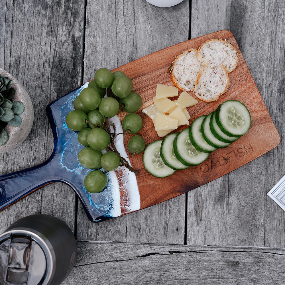 Wooden cutting board with blue resin ocean surf edge and 'Toadfish' branding with sliced cucumbers, cheese, and grapes on a grey wooden surface.