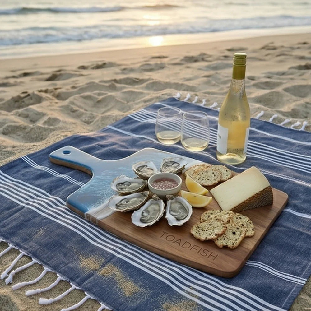 Wood cutting board with blue resin ocean surf edge and 'Toadfish' branding being used in a picnic setup  with oysters, bread, and wine on a blue beach blanket by the ocean.