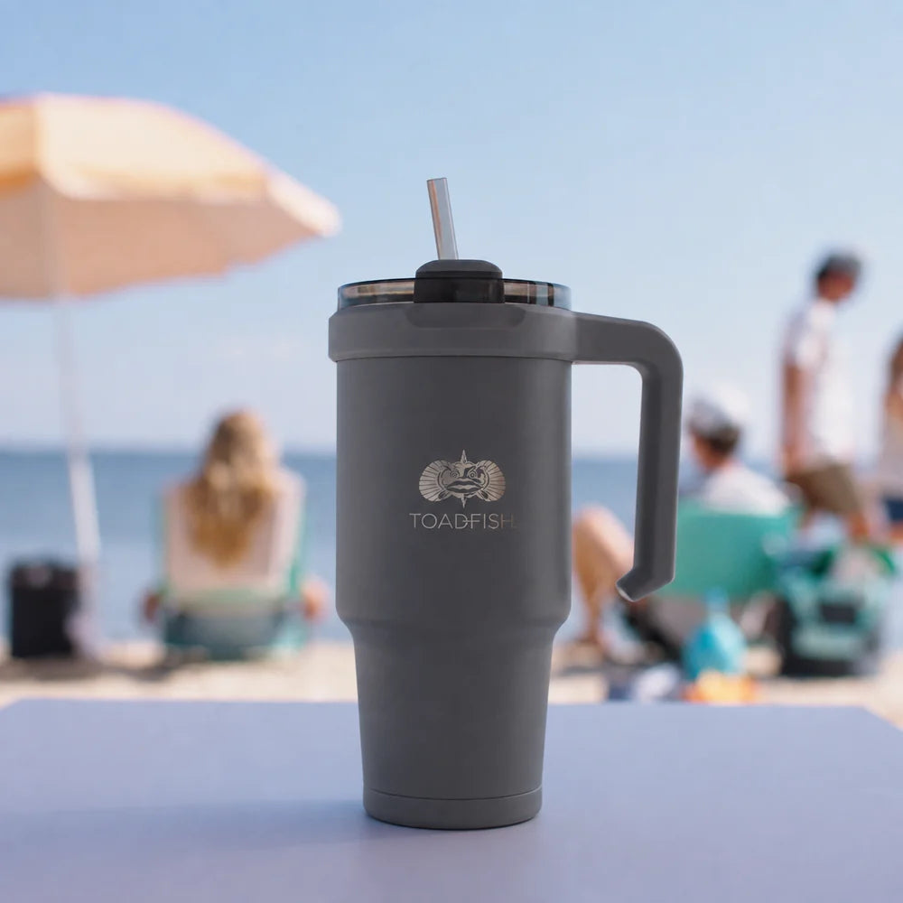 A dark gray insulated travel tumbler with a handle, lid, and straw, standing on a table with a blurred beach scene in the background. The tumbler features the TOABDPH logo printed on the front.