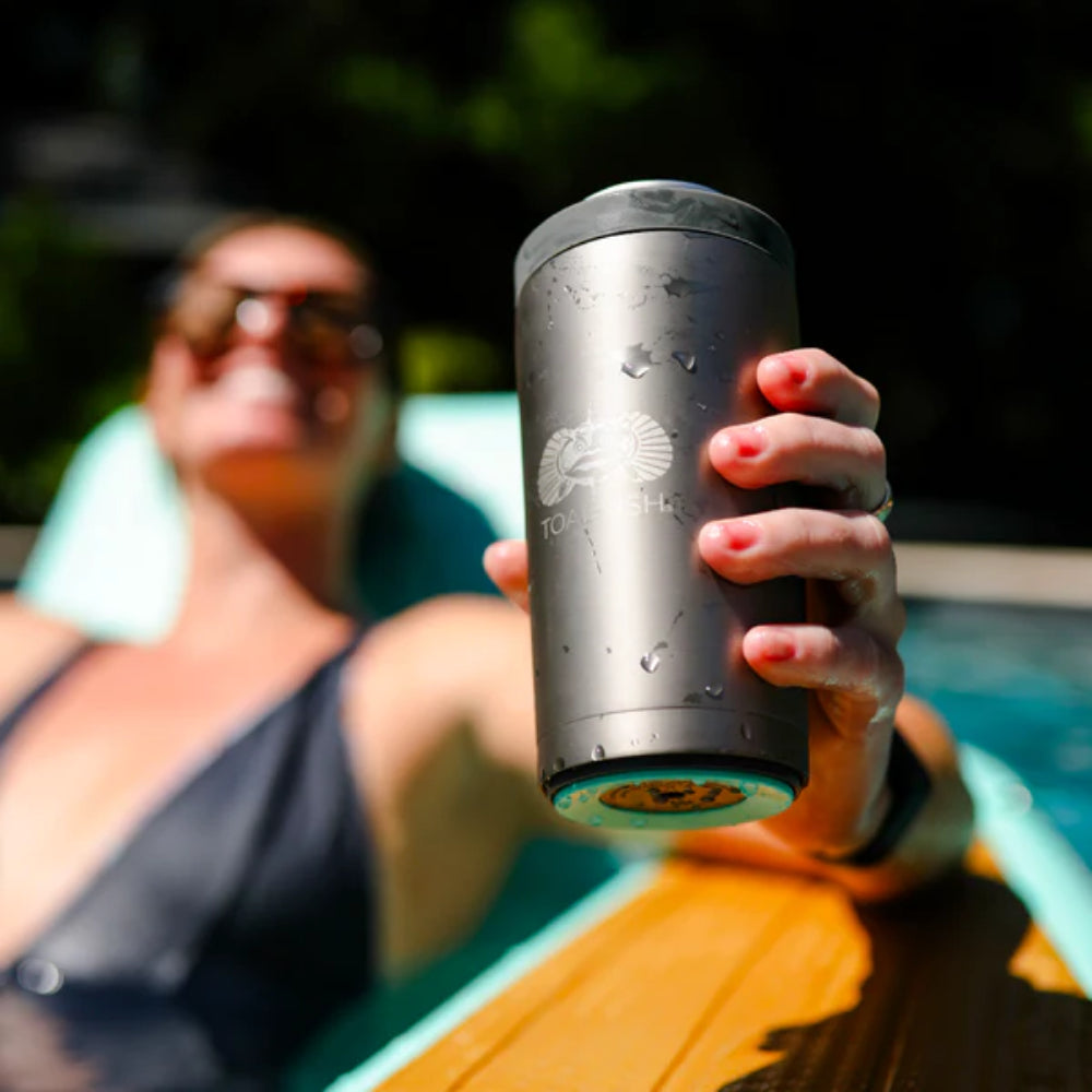 A woman holding a graphite stainless steel universal can cooler with a Toadfish logo on the front next to a pool on a burred backround.