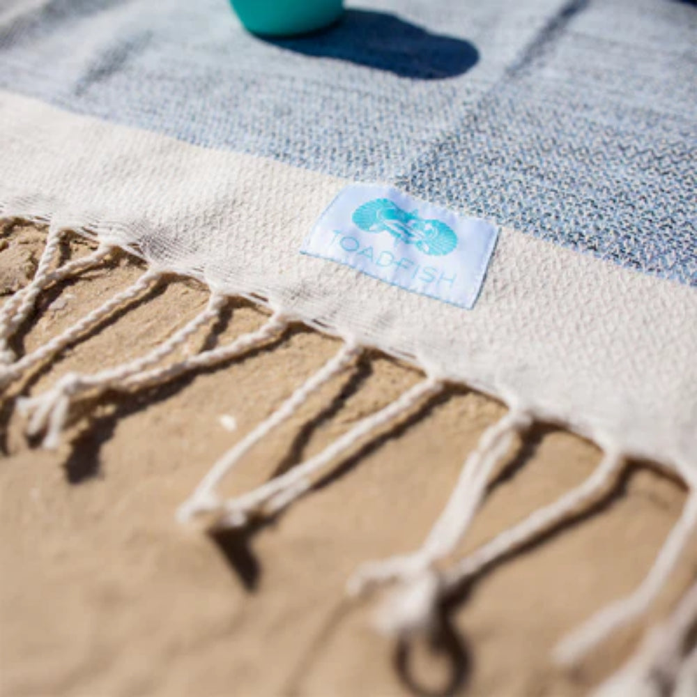 Blue beach towel with a toadfish label and white fringe edge on a sandy surface.
