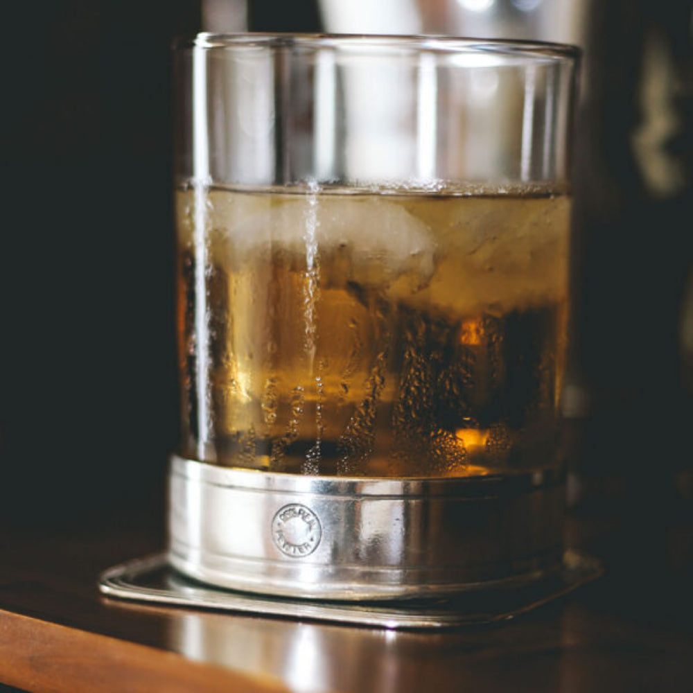 Clear old fashioned glass with silver metallic base with beer in it on a a paper coaster on a table.
