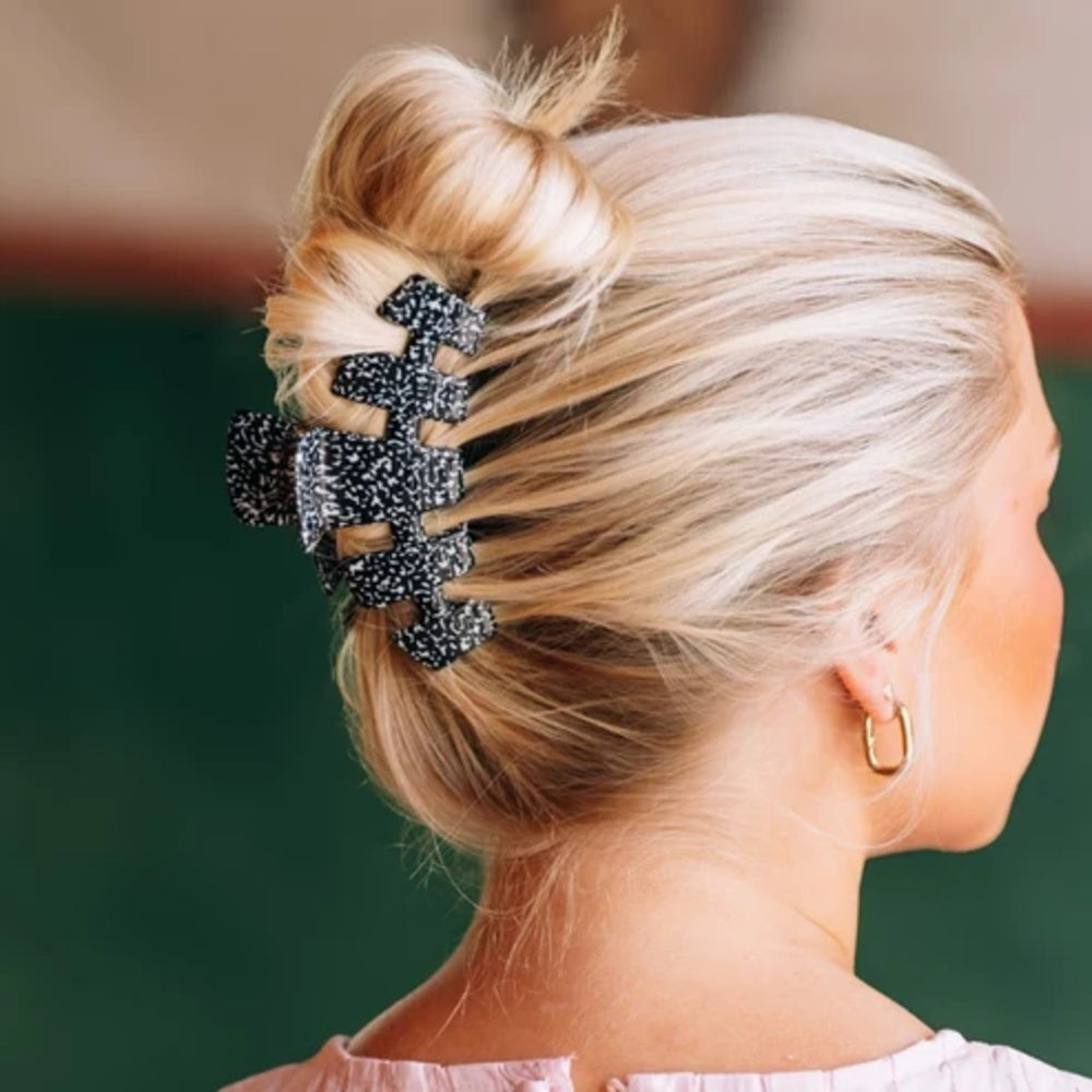 A close-up side view of a woman with blonde hair styled in a messy bun, secured with a large black claw hair clip covered in white rhinestones. She is wearing a small gold hoop earring, and the background is a soft, out-of-focus dark green.