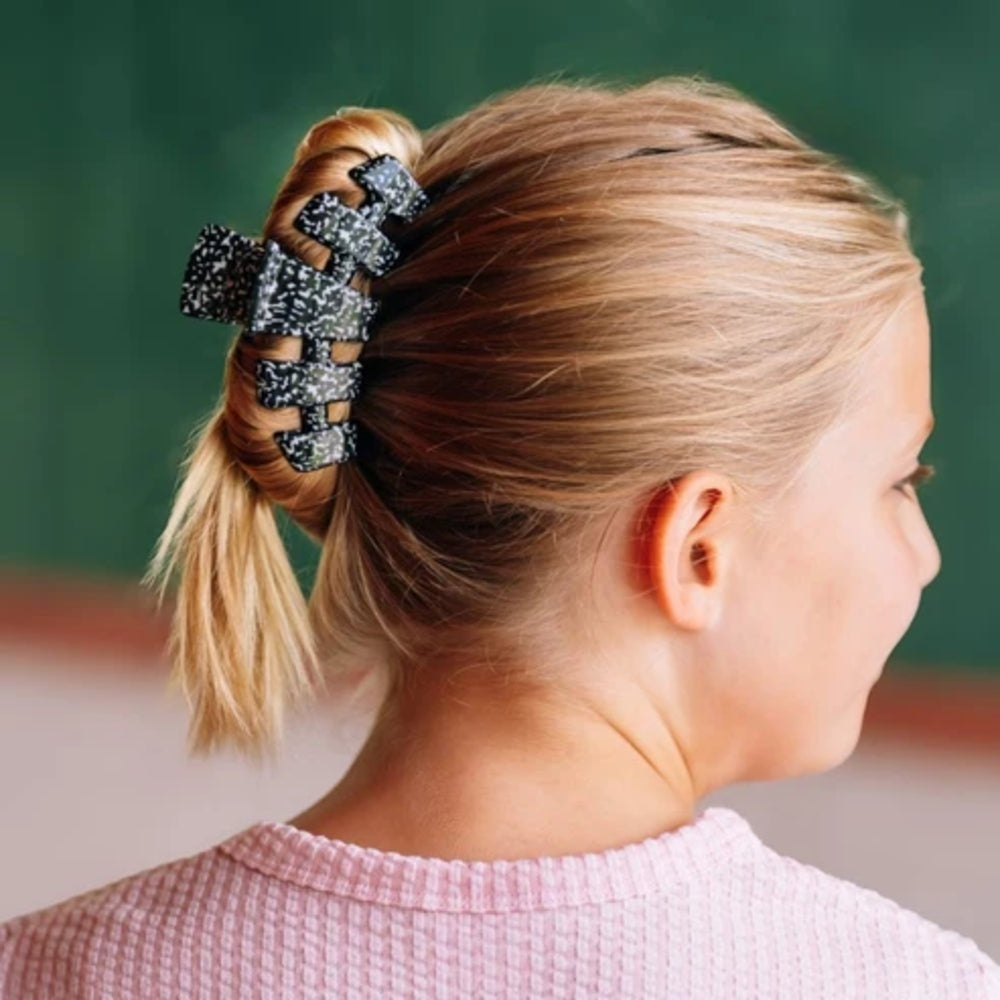 A blonde girl shown from the back and side, wearing a black and white patterned claw clip to hold her hair in a low ponytail. She is wearing a pink textured top against a dark green chalkboard background.