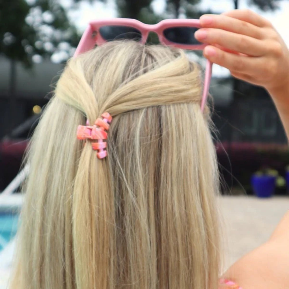 Person with blonde hair wearing pink sunglasses and a pink hair clip outdoors.