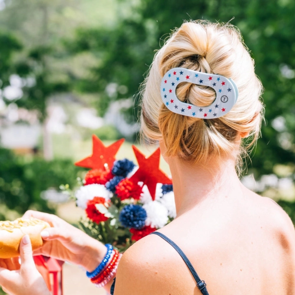 Person with a patriotic hair clip holding a hot dog, with a blurred background of flowers and greenery.