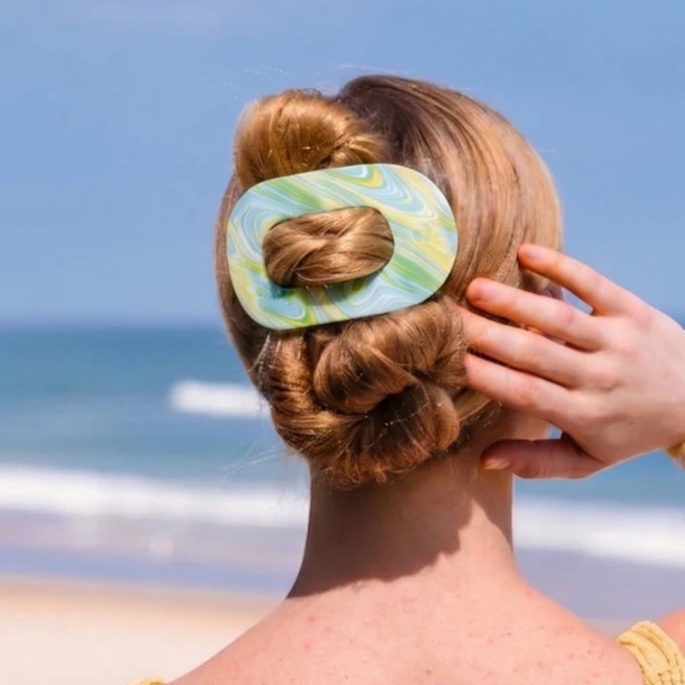 Person with a bun holding a green and blue hair clip against a beach background