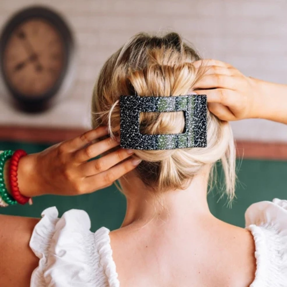 A blonde woman viewed from behind wearing a black glittery rectangular hair claw clip, with her hands adjusting it. She is wearing a white ruffled top, and a green chalkboard and wall clock are visible in the background.