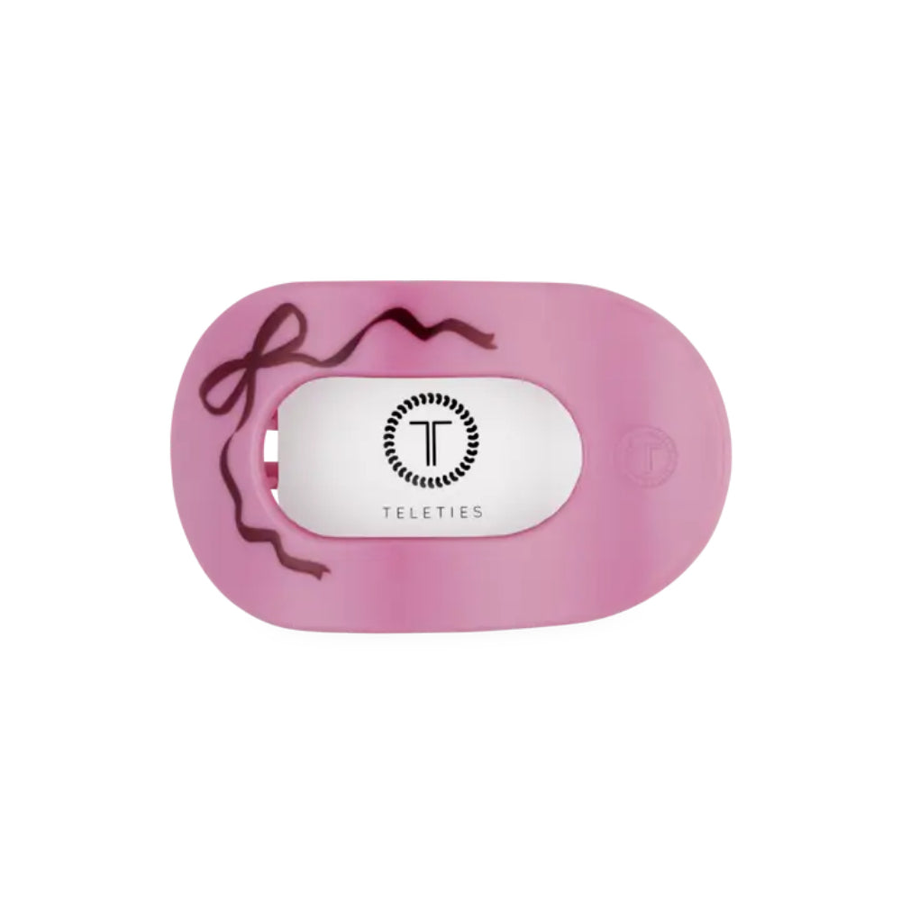 A pink silicone pacifier hair tie with a white central label featuring the Teleties logo and name, decorated with a dark brown ribbon illustration on the left side, shown on a plain white background.