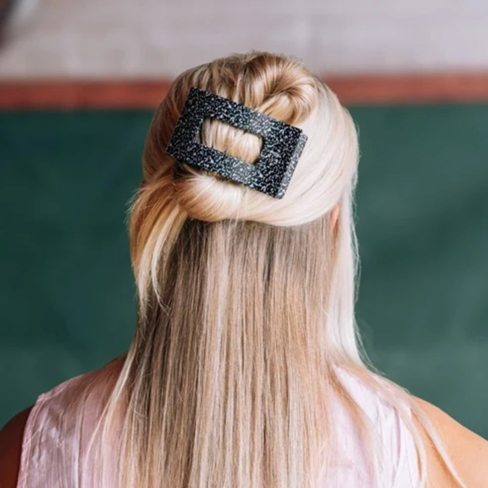 A blonde woman shown from behind wearing a black glittery rectangular hair claw clip in her hair, with a dark green chalkboard background.