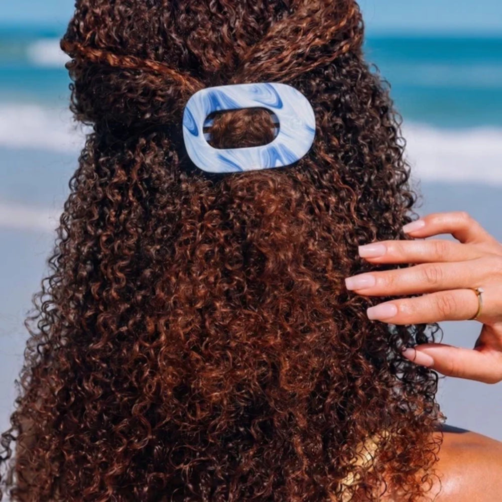 Person with curly hair wearing a blue and white hair clip against a blurred beach background