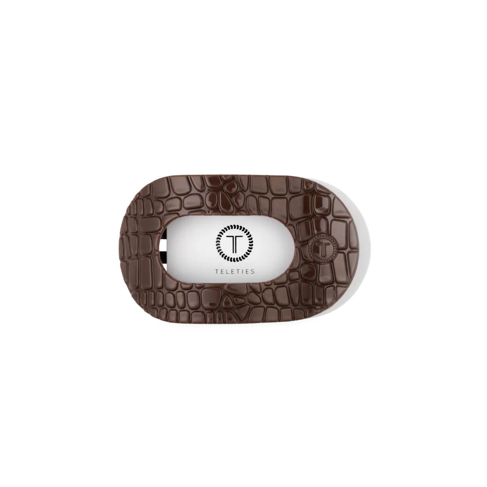 A Teleties travel hair clip case featuring a dark brown crocodile-embossed textured outer shell and a white inner hair clip visible through a central oval cutout.