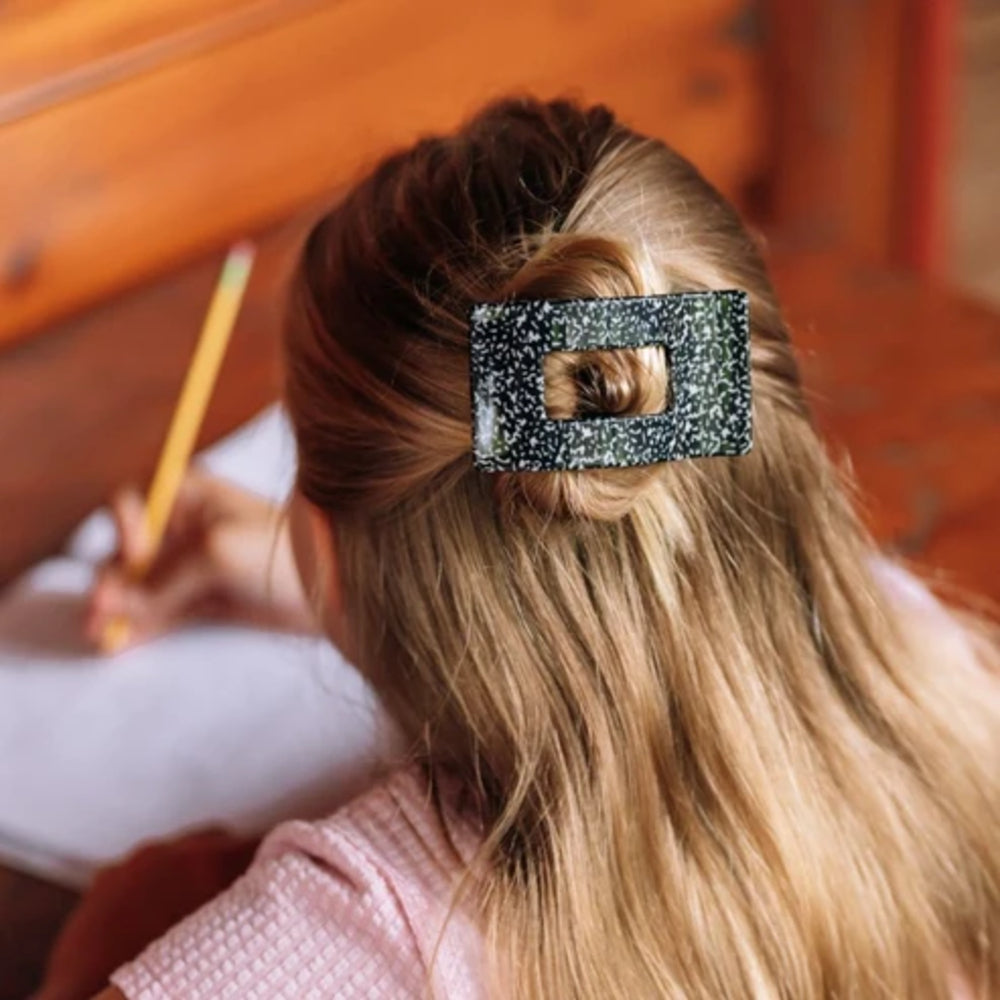 A close-up lifestyle shot of a young girl with long blonde hair seen from behind, wearing a glittery black rectangular hair claw clip. She is writing on a white sheet of paper with a yellow pencil, set against a warm wooden background.