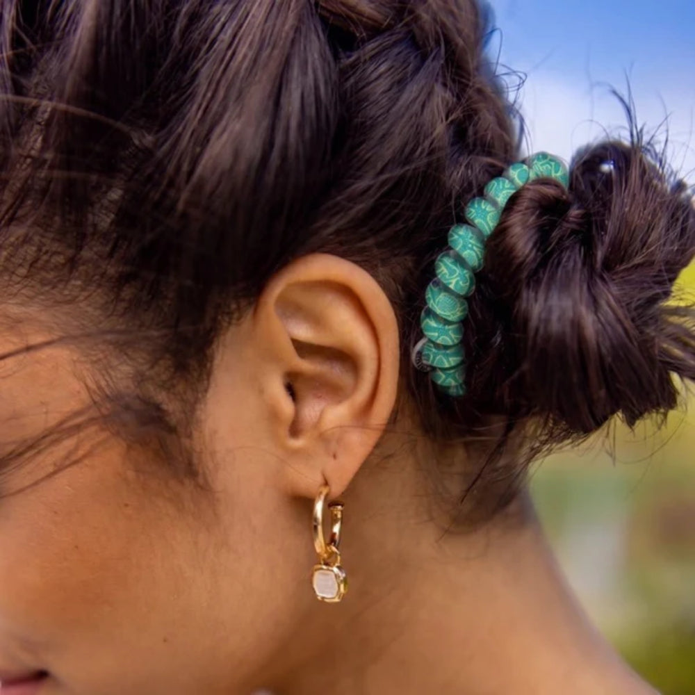Close-up of an ear with a gold earring and a green hair tie in the hair, with a blurred natural background.