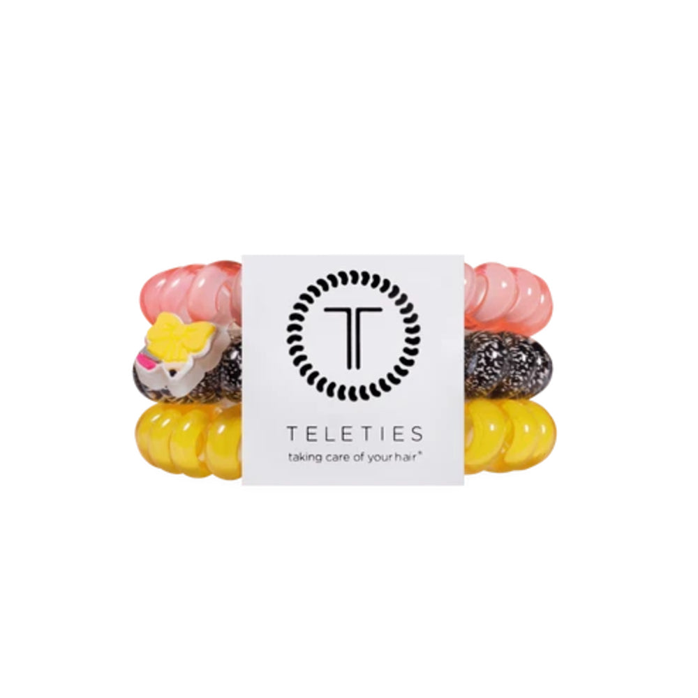 A set of four Teleties hair elastic bands wrapped around a white paper band with the Teleties logo. The bands are translucent pink, yellow, black speckled, and feature a small cow charm.