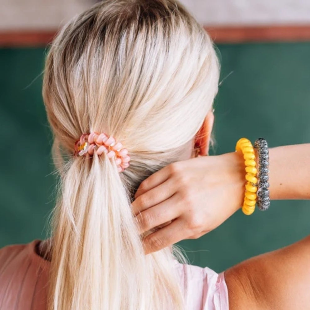 A close-up back view of a woman with blonde hair in a low ponytail, wearing a pink scrunchie. Her right arm is bent in, touching her hair, and she is wearing yellow and gray beaded bracelets on her wrist.
