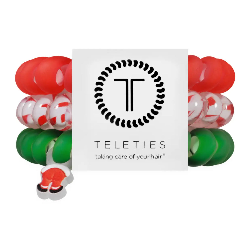 A set of Teleties hair stretch bands arranged around a white brand card. The bands are red, green, and clear with red patterns, and a small Santa hat charm is attached on the left.