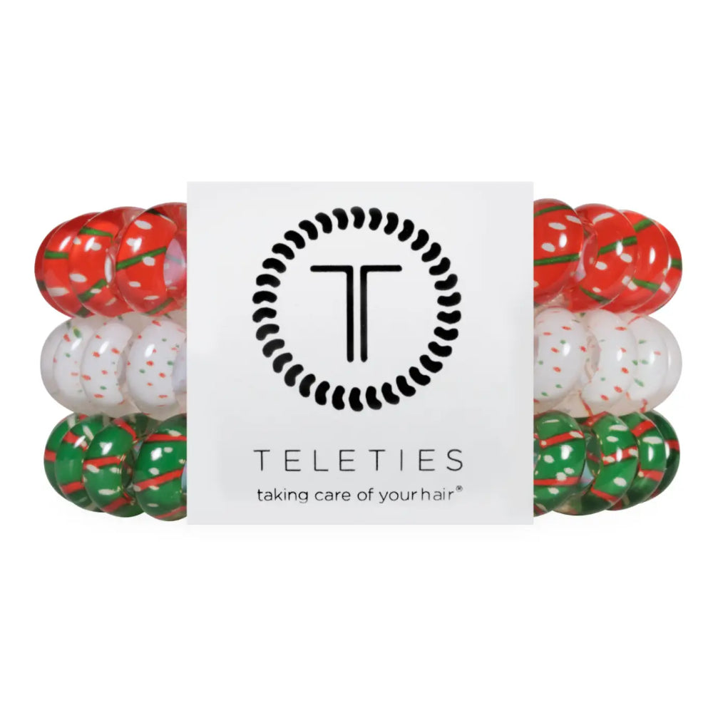 Three elastic hair extension bands in red, white, and green holiday patterns, arranged symmetrically on either side of a white central card with the Teleties logo.