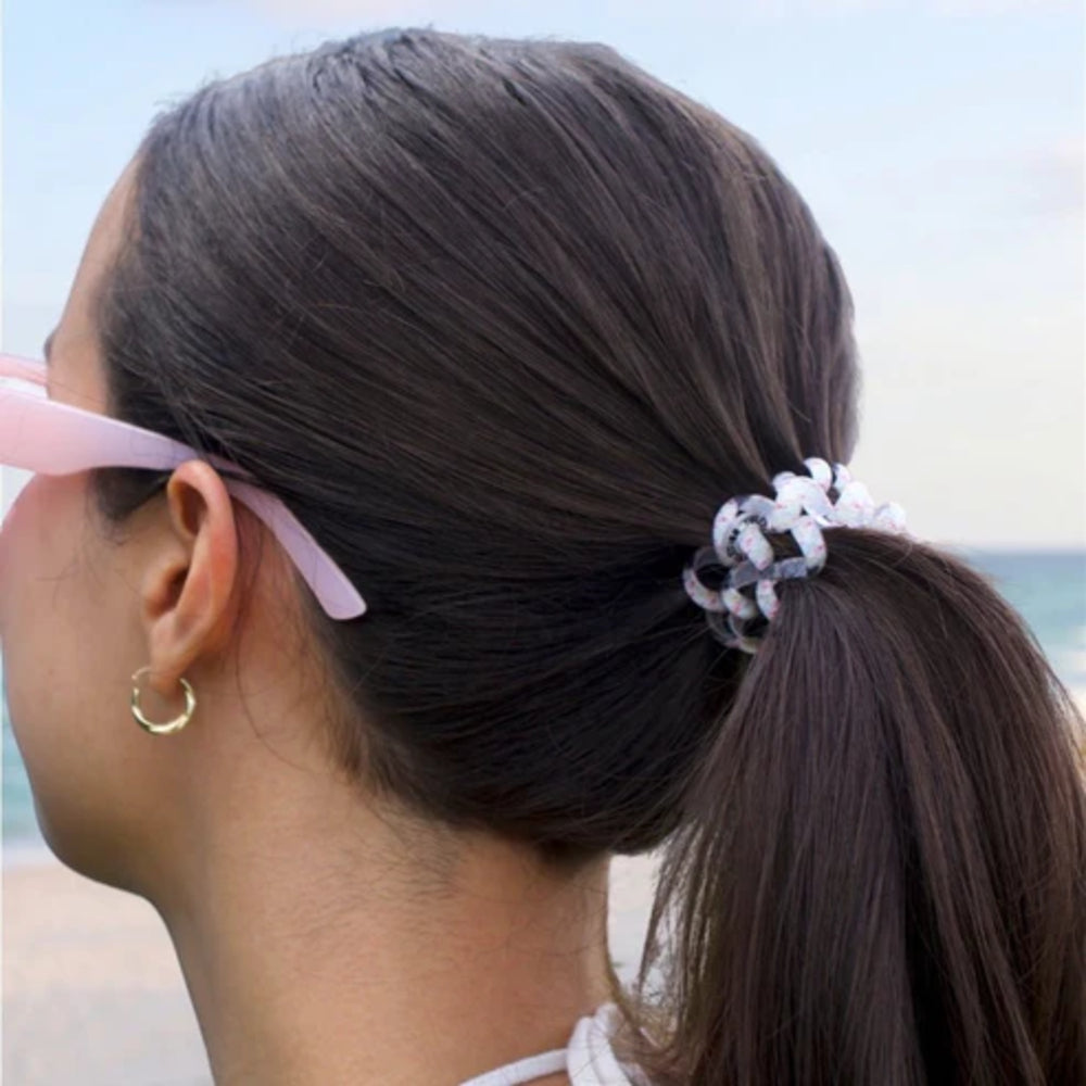 Person with a ponytail wearing pink sunglasses, hair ties, standing by a beach.