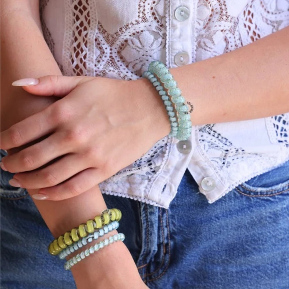 A close-up of a person wearing multiple beaded bracelets on both wrists. The bracelets include light mint green, pale yellow, and light blue-green beads, with one bracelet featuring a small gray stone accent.