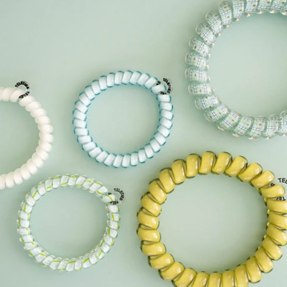 Five coiled stretch bracelets arranged on a pale mint green background. The bracelets are translucent and come in white, aqua, yellow, and clear-and-white patterns, each with a small black-and-white logo tag.