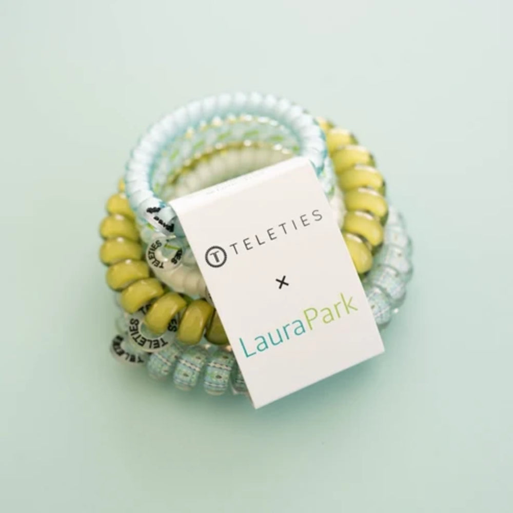 A stack of coiled hair extension bands in pastel yellow, white, and light blue, with a white paper tag reading 'TELETRIES x LauraPark' placed over the top.