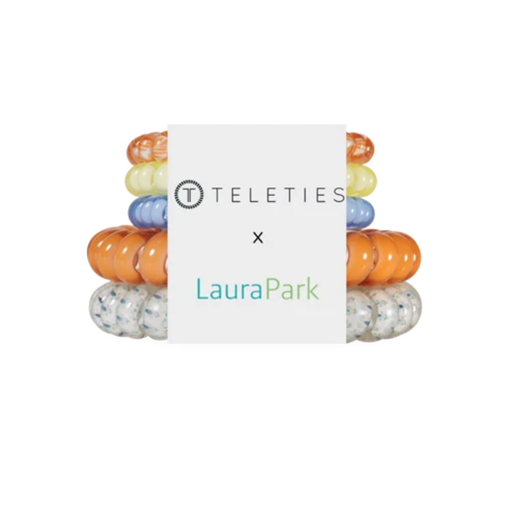A pack of Teleties x LauraPark silicone baby teething rings, featuring translucent orange, pale yellow, blue, and speckled clear rings, wrapped in a white paper band with the brand logos.