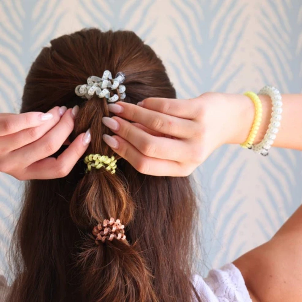 Three pearl-bead hair scrunchies in white, yellow, and pink are shown on a model's brown hair, with hands adjusting the top scrunchie.