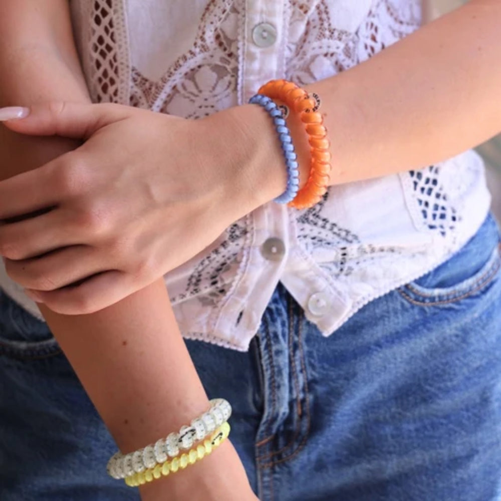 A close-up of a person's wrists wearing three colorful beaded stretch bracelets: a white and light blue bracelet, an orange bracelet, and a yellow bracelet, styled with a white lace top and blue jeans.
