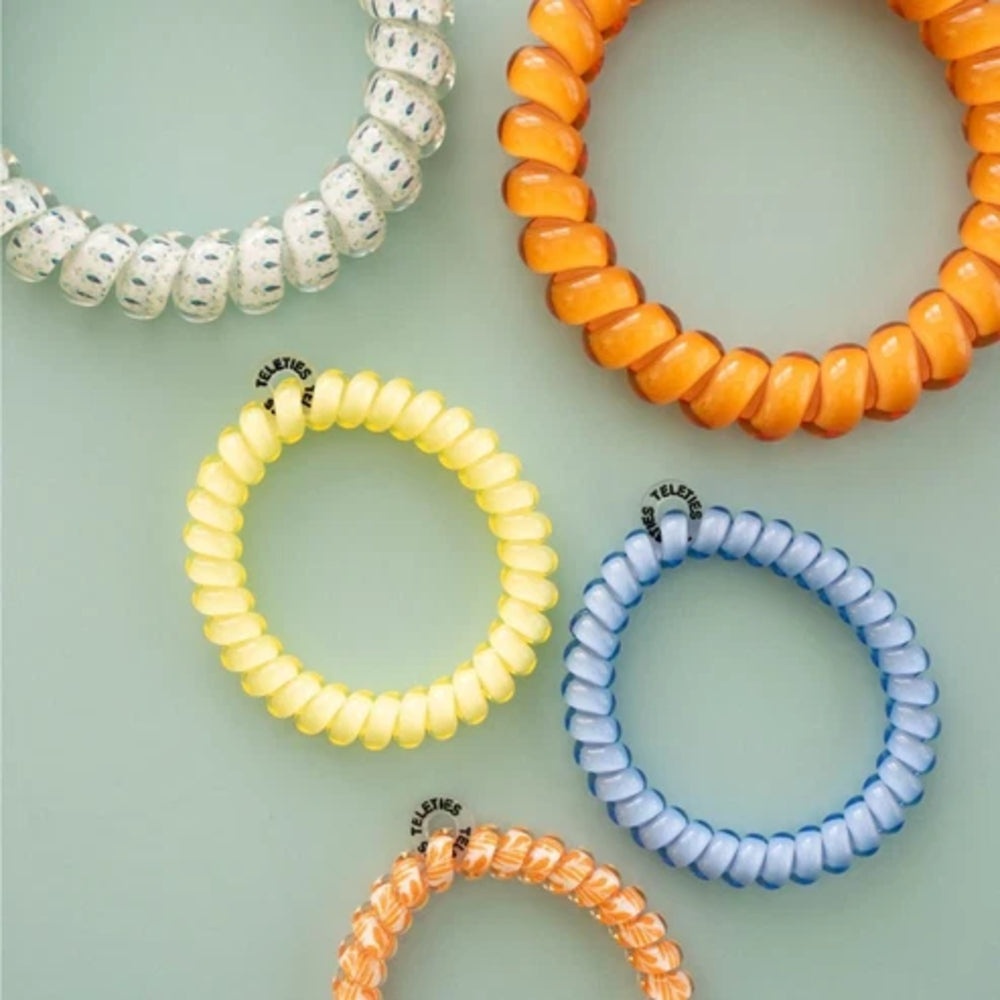 Four coiled elastic hair extension bands in white, orange, yellow, and light blue, arranged on a pale mint green background. Each band has a small black metal clasp with the brand name 'FLEXY' printed on it.