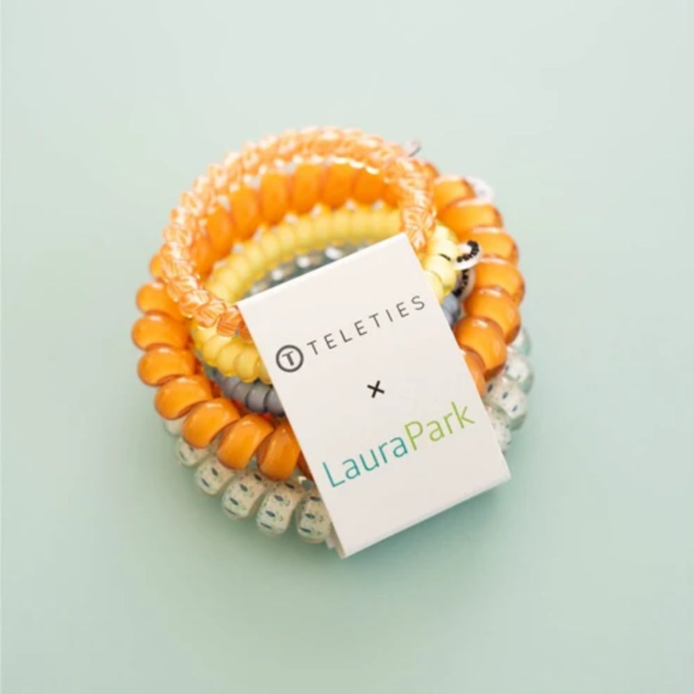A set of coiled hair extension bands in orange, yellow, gray, and white, wrapped with a white paper tag that reads 'TELETIES x LauraPark' on a pale mint background.