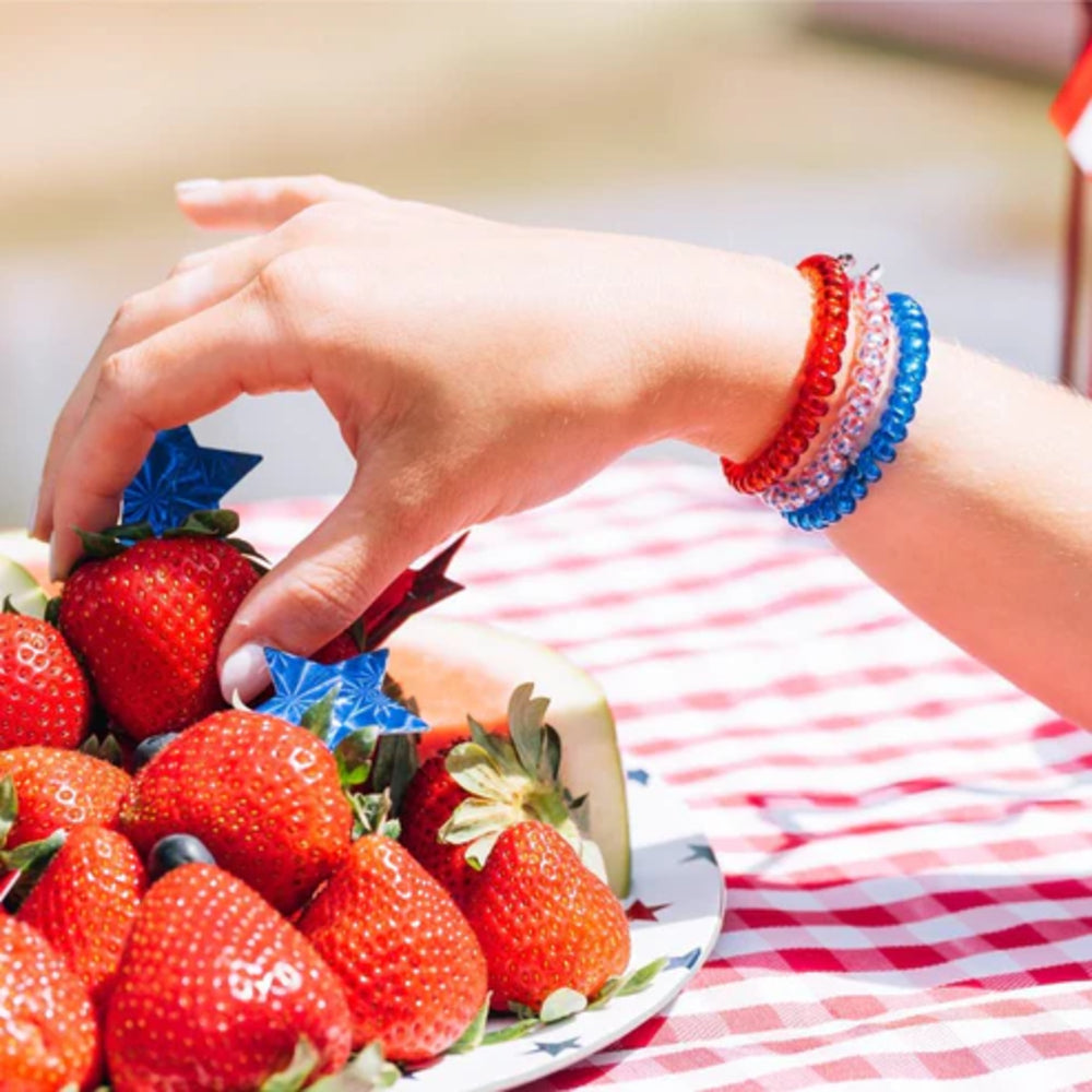 Hand picking strawberries from a plate with a checkered tablecloth and patriotic bracelets.