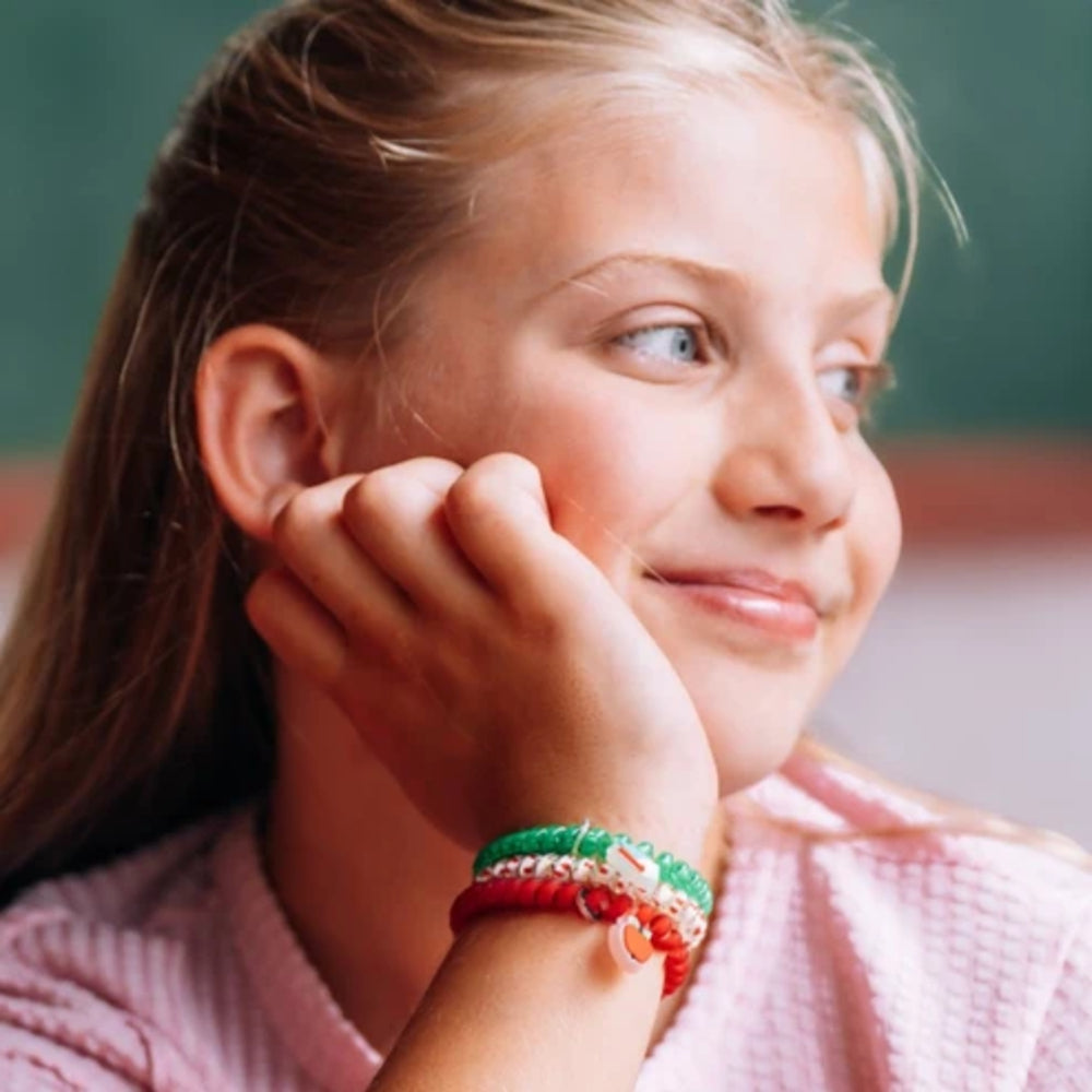 A close-up lifestyle shot of a young girl with light brown hair resting her chin on her hand, wearing a set of colorful braided bracelets on her wrist. She is wearing a pink textured shirt against a soft, blurred green background.