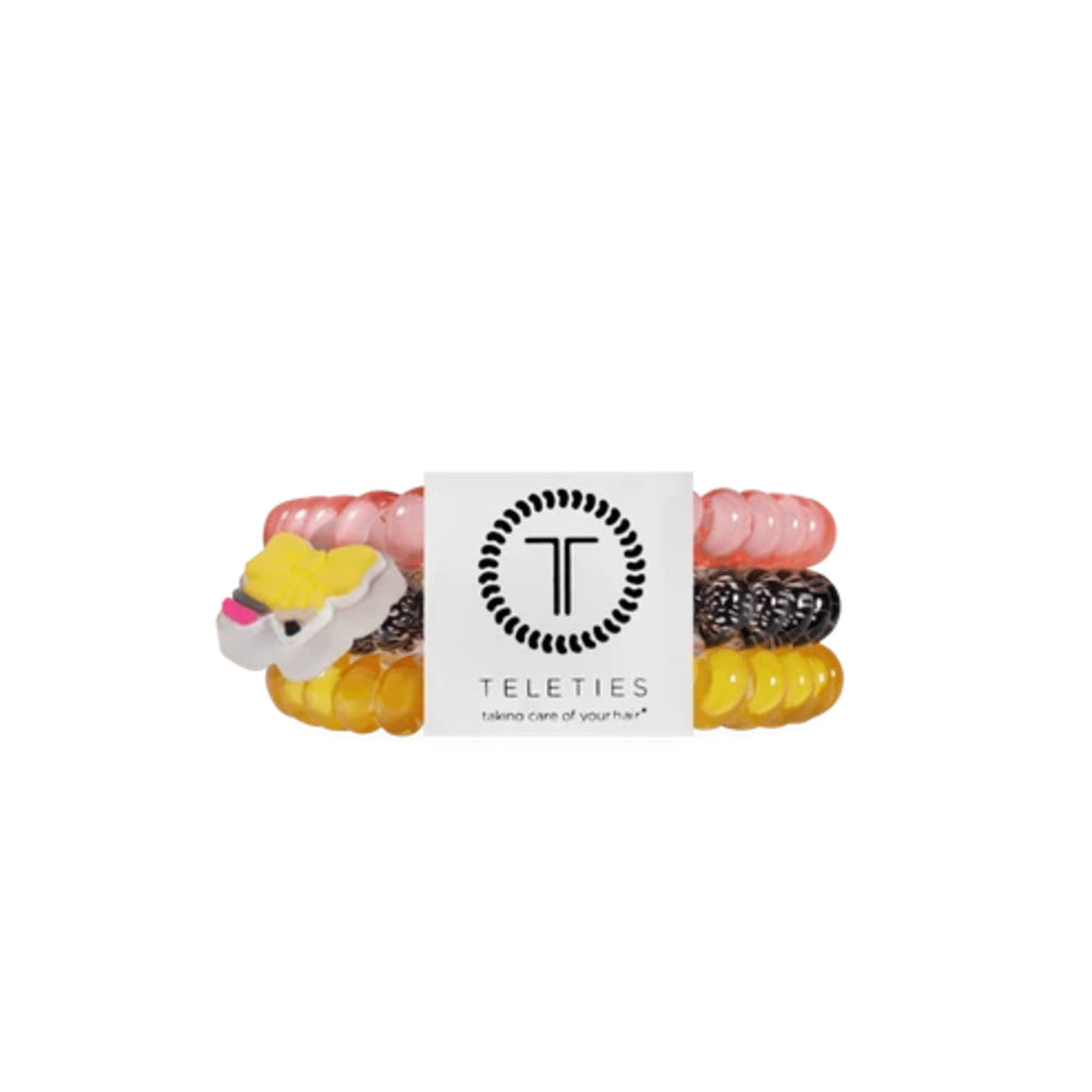 A set of four Teleties hair elastic bands in pink, black-and-white speckled, and yellow, with a small white and yellow cow charm, wrapped in a white paper band featuring the Teleties logo.