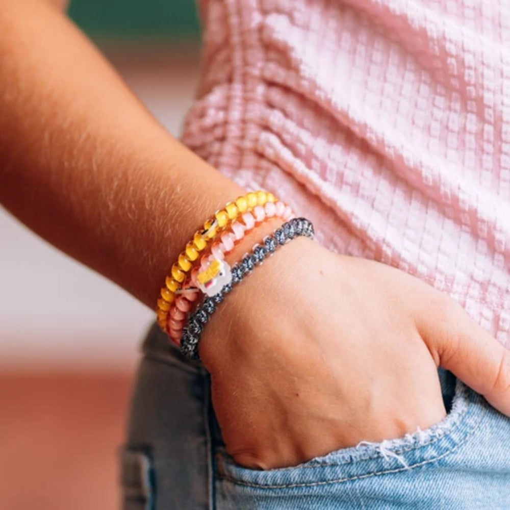 A close-up of a person's hand in a denim pocket wearing three beaded bracelets: yellow, light pink, peach, and dark gray with a small white flower charm.