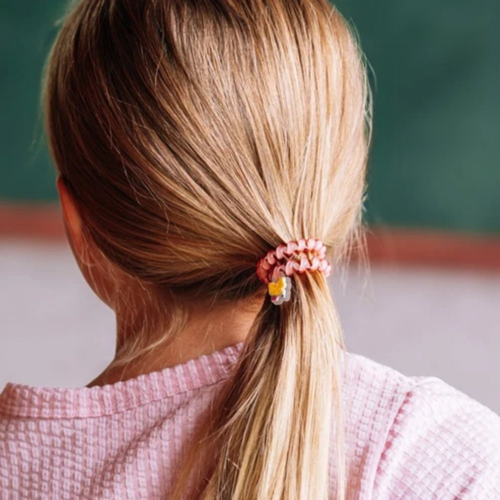 A close-up back view of a young girl with blonde hair tied in a ponytail with a pink textured scrunchie and a small yellow charm, wearing a pink waffle-knit top.