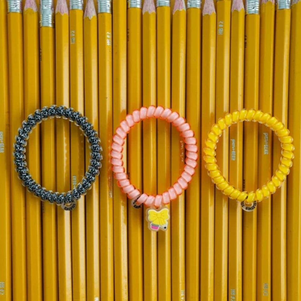 Three coiled elastic hair ties in black, pink, and yellow are arranged in a row on a background of sharpened yellow pencils. The center pink tie has a small yellow bow charm.