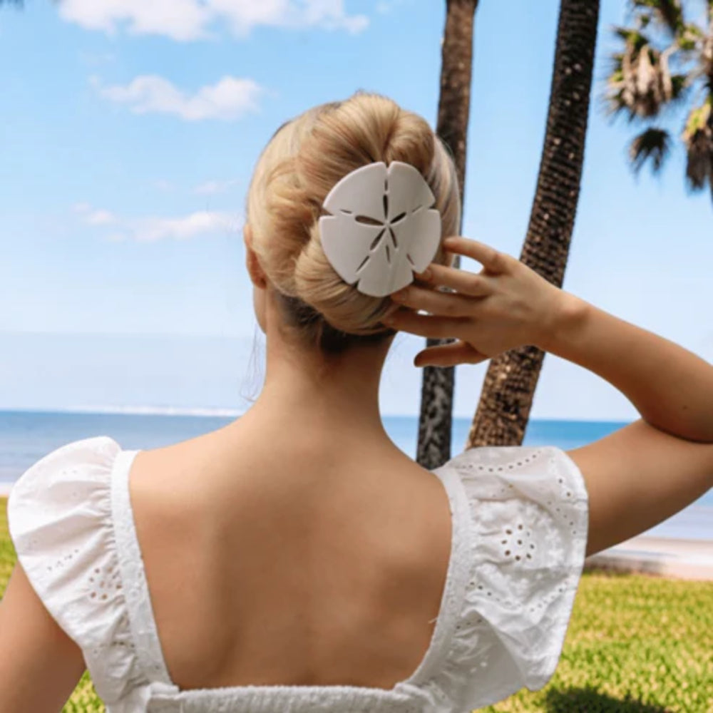 Woman holding a sand dollar hair clip against her hair with palm trees and ocean in the background