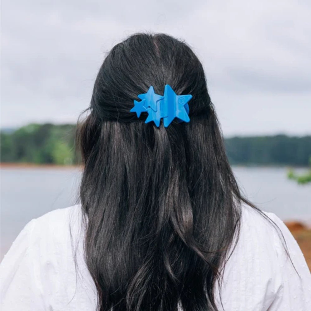 Person with blue hair clip in their dark hair, standing outdoors with a lake and trees in the background.