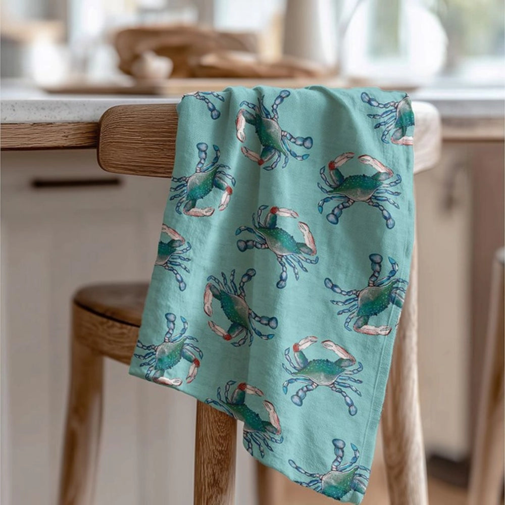 A light aqua kitchen towel with a repeating blue and orange crab pattern, draped over the back of a wooden chair in a kitchen setting.