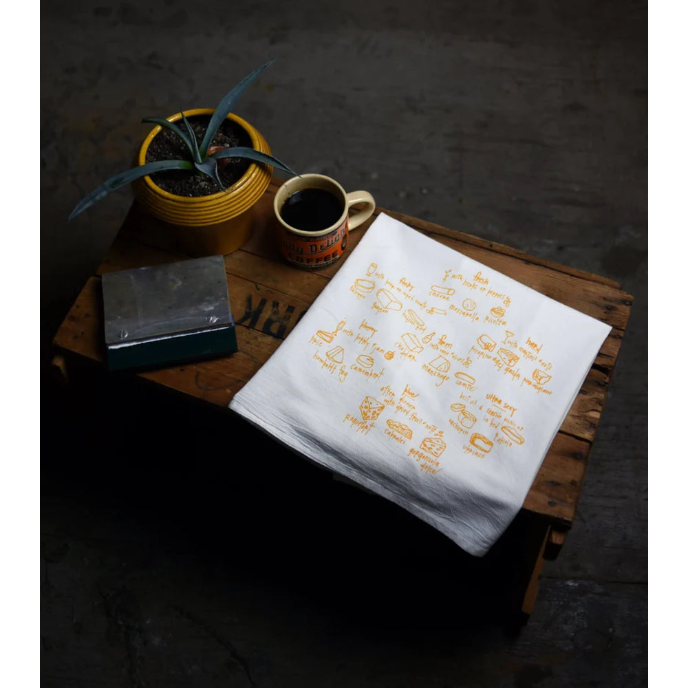 White towel with gold text and graphics on a wooden surface with a cup of coffee and plant.